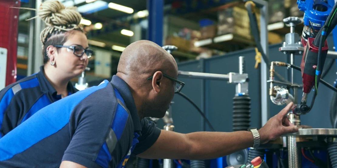 Two people in blue ERIKS shirts at an assembly cell. One leans in with a hand on a part under the tooling. The other stands behind him, watching the same point.