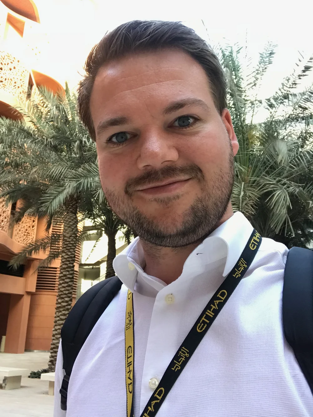 Max Redecker outside the Etihad Airways offices in Abu Dhabi, wearing an Etihad lanyard, palm trees and the building's perforated facade behind him.