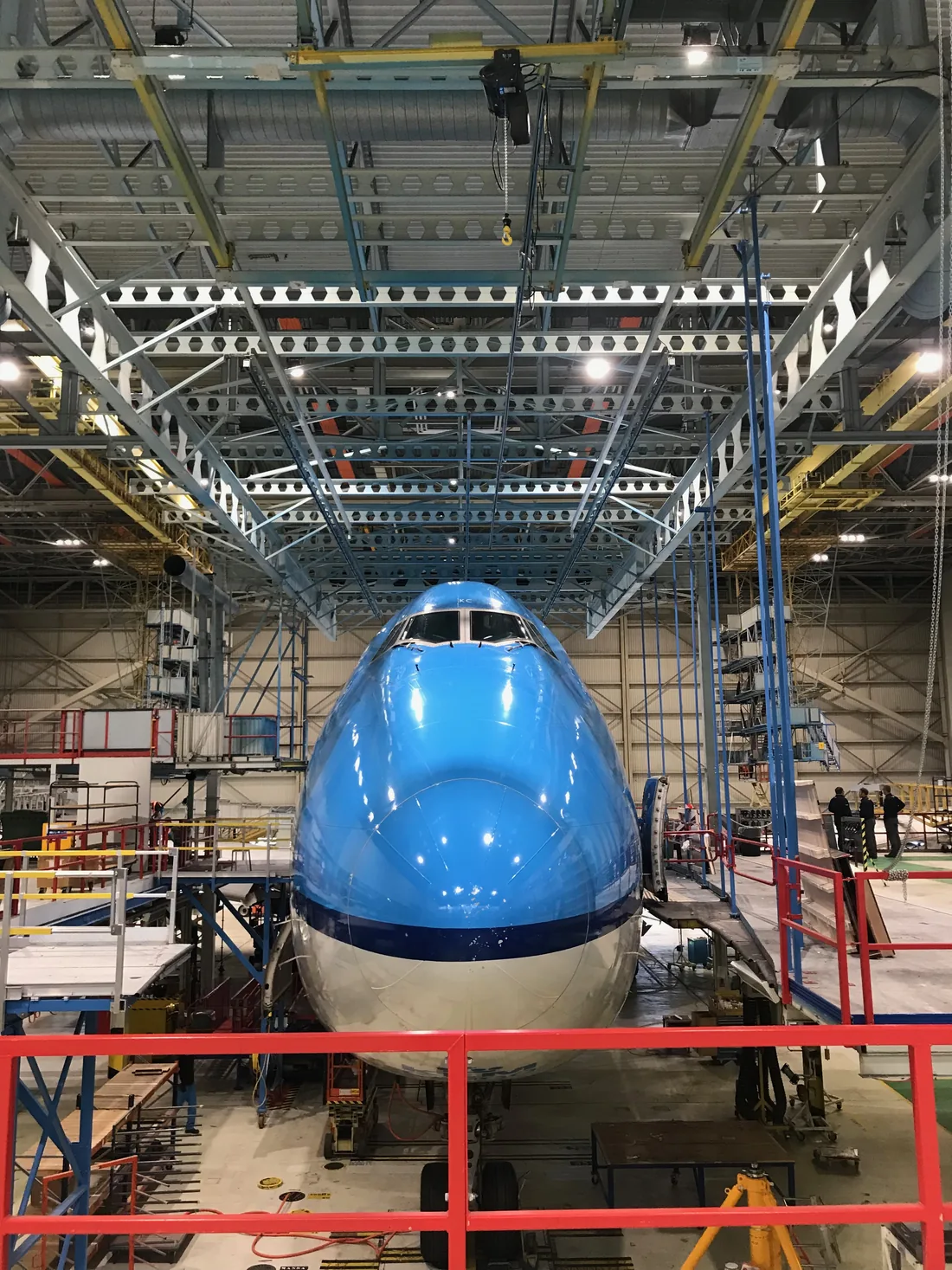 The nose of a KLM Boeing 747 head-on inside the hangar, enclosed by scaffolding and a red work platform.