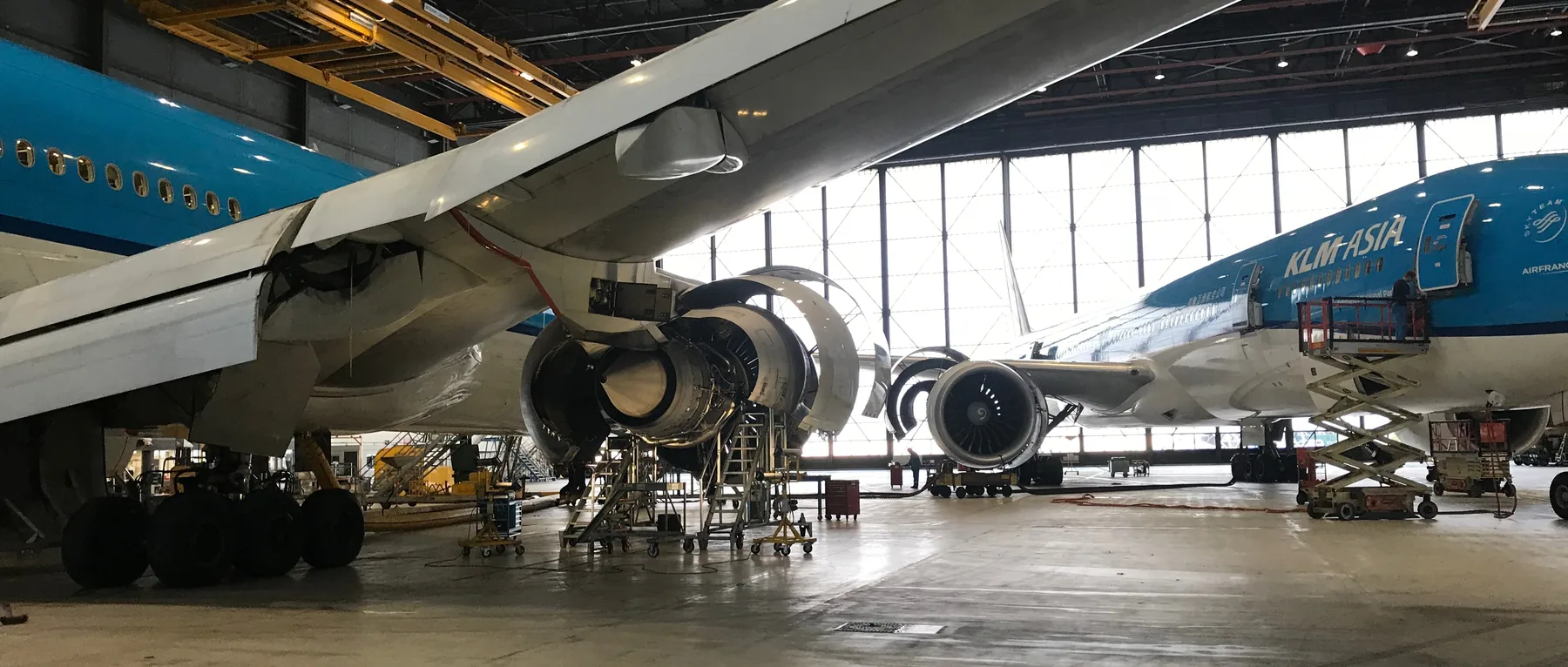 Two KLM aircraft in the maintenance hangar, one with its engine cowlings open over a row of work platforms.